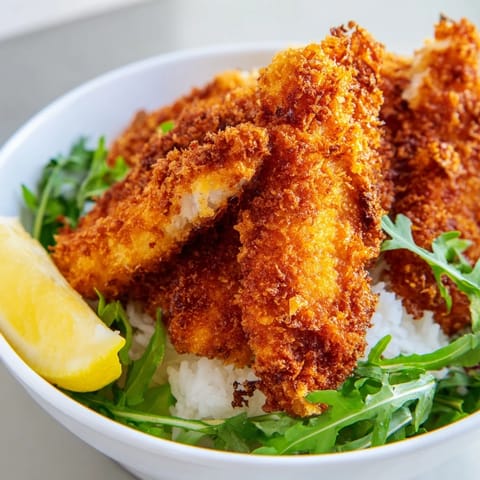 A close-up of golden-brown, crispy Parmesan chicken cutlets resting on steamed rice beside a bright arugula salad.